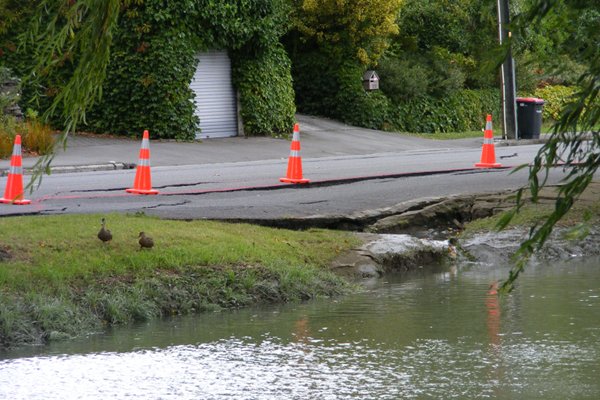A road running along a riverbank which has collapsed following an earthquake. There are cones along the road to mark the danger, and ducks on the riverbank.
